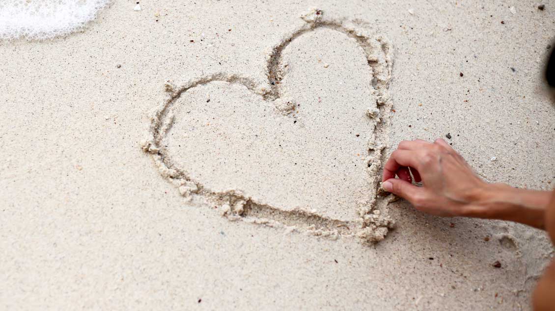 Ein Finger zeichnet ein Herz in den Sand.