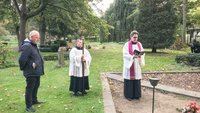 Drei Personen stehen auf einem Friedhof. Link im Bild steht ein schwarz gekleideter Mann. In der Mitte steht ein Messdiener mit Kreuz in der Hand. Rechts steht ein Pfarrrer in Gewand mit Buch in der Hand.