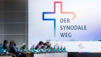 An einer Wand steht ein großes Logo in Form eines bunten Kreuzes. Daneben steht der Text "Der Synodale Weg"