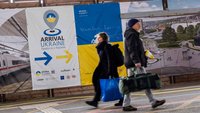 Zwei Menschen, die auf einem Bahnsteig laufen. Im Hintergrund eine Ukraine-Flagge.