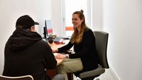 Ein schwarz gekleideter Mann sitzt auf einem Stuhl mit dem Rücken zur Kamera zugewandt. Eine jüngere Frau schaut ihn freundlich an. Sie hat einen Stift in der Hand.