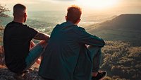 Zwei junge Männer, von hinten fotografiert, sitzen auf einem Berg und schauen in die Ferne