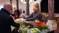 Eine Frau mit einer orangenen Schürze reicht einem Mann einen Salatkopf