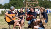 Kinder sitzen auf einer Bank und hören einem Gitarrenspieler zu.