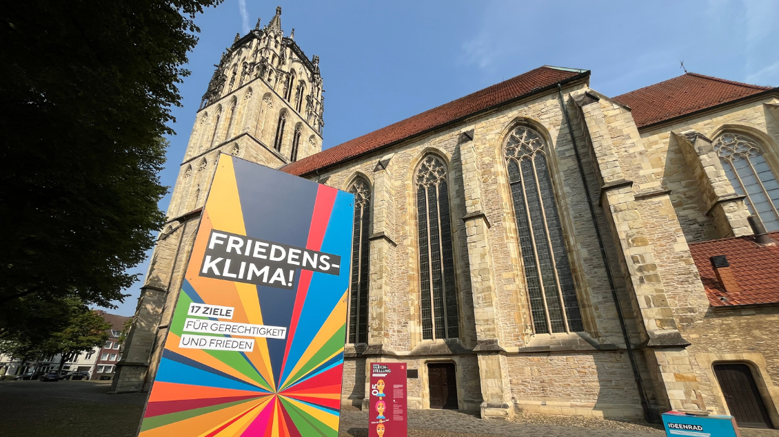 Bunte Stele mit der Aufschrift "Friedensklima! 17 Ziele für Gerechtigkeit und Frieden" steht vor der Liebfrauen-Überwasserkirche in Münster.